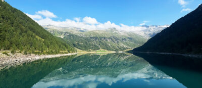 ein bergsee in den alpen mit bergpanorama im hintergrund und hellblauem himmel mood bild für alpen kollektion von jan spille schmuck