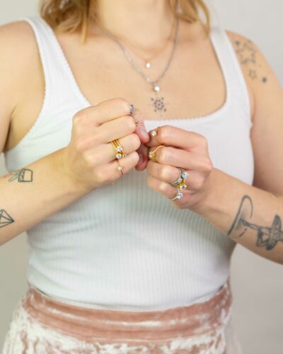 Dekoltée und Fäuste einer Frau mit weißem Shirt zeigen Ringe und Ketten aus der Chaton Kollektion mit Krappenfassung von Jan Spille Schmuck vom Goldschmied